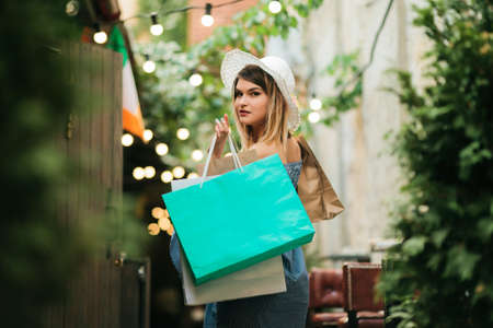 Shopping concept. Young attractive woman in a hat and dress holds shopping bags in her hands while walking along the street. Back viewの写真素材