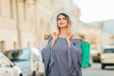 Happy shopping concept. Young attractive woman in a hat and dress holds shopping bags in her hands and smiles in a summer cityの写真素材