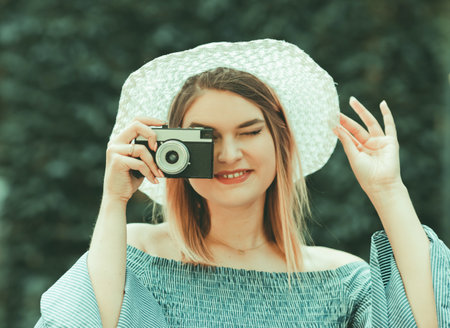 Vintage portrait. Young spectacular woman in dress and hat takes pictures with retro camera on blurred outdoor backgroundの写真素材