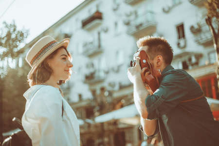 Young cheerful couple of hipsters are photographed on a retro camera while walking around the city. Male photographer photographs a stylishly dressed young womanの写真素材