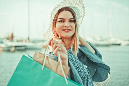 Portrait of a cheerful young shopaholic woman in a hat and dress on a background of the sea. Happy shopping concept.の写真素材