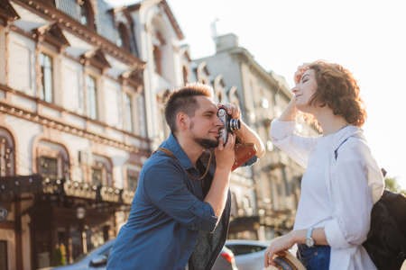 Young cheerful couple of hipsters are photographed on a retro camera while walking around the city. Male photographer photographs a stylishly dressed young womanの写真素材