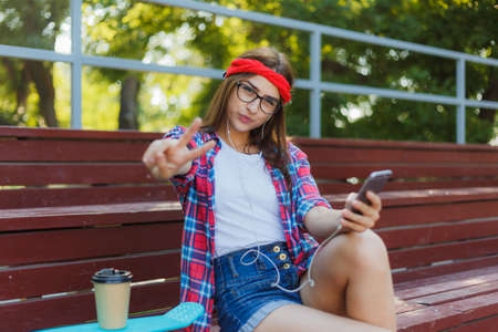 Young woman skater dressed in stylish clothes sits in the stands in a skatepark holds a smartphone in her hands and listens to music in earphones on a bright sunny dayの写真素材