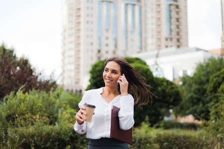 Businesswoman. Young attractive female manager wearing a skirt and a blouse holding coffee cup and talking on phone at background of the city landscape. All in business, coffee on the goの写真素材