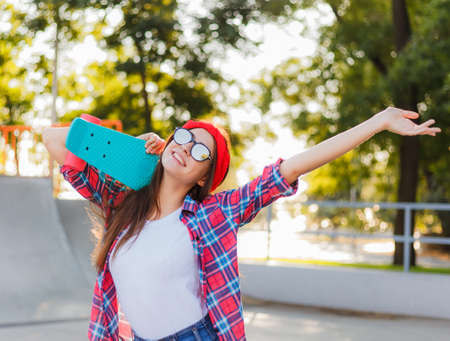 Cheerful funny young woman dressed in stylish clothesand mirrored sunglasses while holds skateboard in her hand in skatepark at bright sunny day. Summer funの写真素材