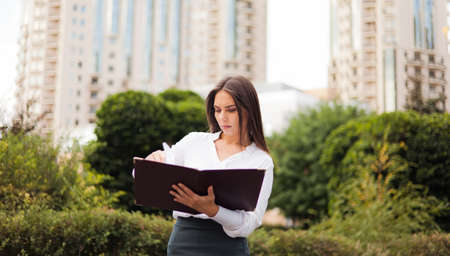 Businesswoman. Young attractive female manager wearing a skirt and a blouse holding folder with documents at background of the city landscape. All in businessの写真素材