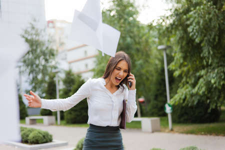 Brought to hysteria, angry business woman dressed in a white blouse and skirt, screaming in the phone, throws out sheets of paper outdoors in the cityの写真素材
