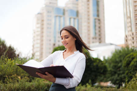 Businesswoman. Young attractive female manager wearing a skirt and a blouse holding folder with documents at background of the city landscape. All in businessの写真素材