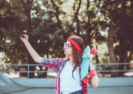 Funny young woman in glasses and red plaid shirt. Hipster girl n skatepark while makes selfie with skateboard in skatepark at sunny bright day. Summer funの写真素材