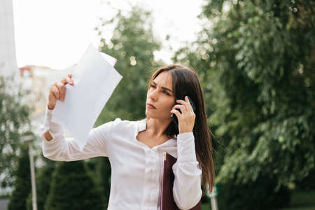 Young attractive female manager holds sheets of paper talking on the phone outdoors in the city. All in businessの写真素材