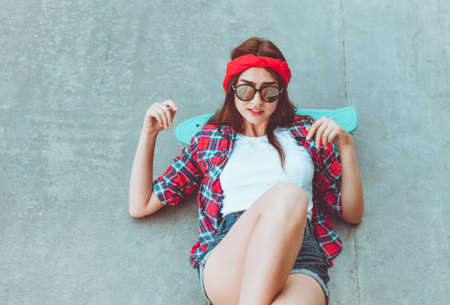 Young hipster girl in stylish mirrored glasses, shorts, red plaid shirt and bandage on her head lies with skateboard on concrete surface in skateparkの写真素材