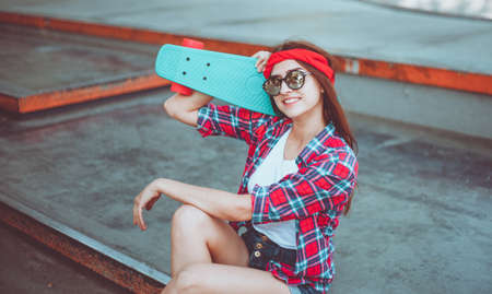 Portrait of stylish skater woman in skatepark. Attractive girl in red plaid shirt, mirrored sunglasses, shorts rests with a skate. Summer lifestyle image of trendy pretty young girlの写真素材