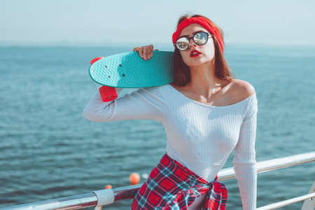 Portrait of young stylish woman in fashionable outfit with skate in hands on beach against sea background. Summer lifestyle image of trendy pretty young girlの写真素材