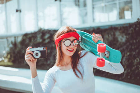 Retro style. 80s. Portrait of stylish dressed young woman with skateboard and film camera outdoors on bright sunny day. Summer lifestyle image of trendy pretty young girlの写真素材