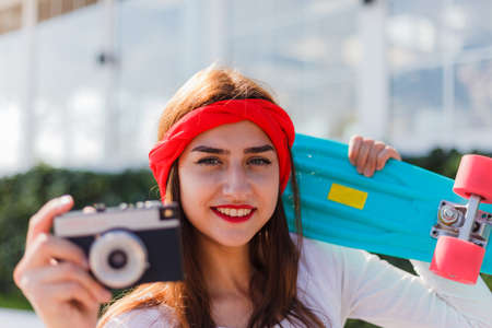 Retro style. 80s. Portrait of stylish dressed young woman with skateboard and film camera outdoors on bright sunny day. Summer lifestyle image of trendy pretty young girlの写真素材