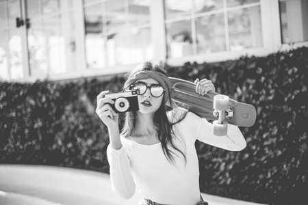 Retro style. 80s. Portrait of stylish dressed young woman with skateboard and film camera outdoors on bright sunny day. Summer lifestyle image of trendy pretty young girlの写真素材