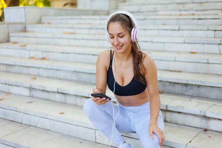 Concept of rest after training. Young happy sport woman in sportswear use smartphone and listens to music in headphones while sitting on stairs on bright sunny dayの写真素材