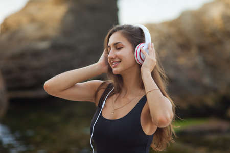 The concept of relaxing on the beach. Summer rest. Young sexy woman in a swimsuit listens to music with headphones on a wild beach with stonesの写真素材