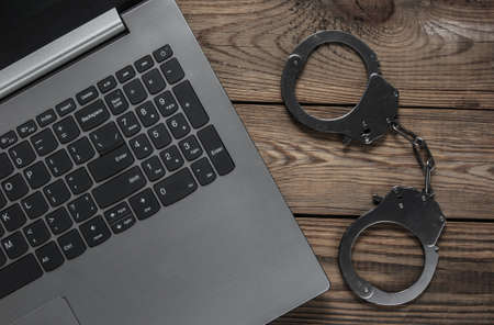 Cybercrime, online digital theft. Laptop with steel handcuffs on wooden table. Top viewの写真素材