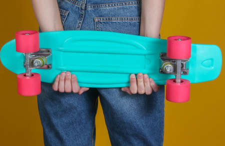 Young slim woman in jeans holds plastic cruiser board in her hands on yellow background. Youth...