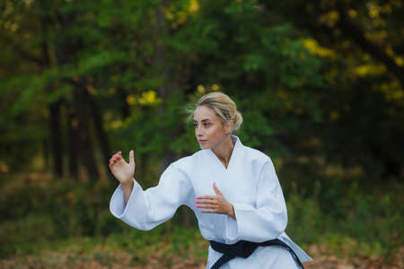 Attractive female master fighter in a white kimono with a black belt stands in a fighting stance outdoorsの写真素材