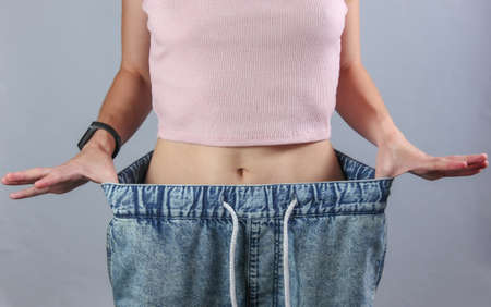 The concept of losing weight. Woman in very big jeans on gray studio background. Crop photo