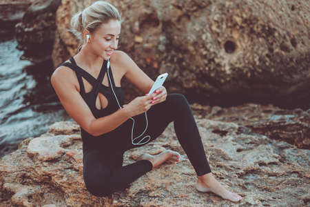 Young slim fit woman in sportwear listening to music in earphones and using smartphone while sitting on a stone at wild beachの写真素材