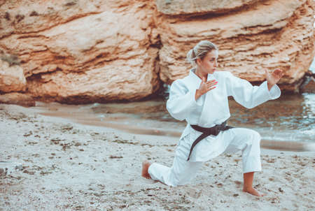Attractive female martial artist in a white kimono with a black belt stands in fighting stance on wild beachの写真素材