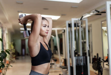 Slim fit woman in sportswear doing extension exercise from behind the head with dumbbells in her...