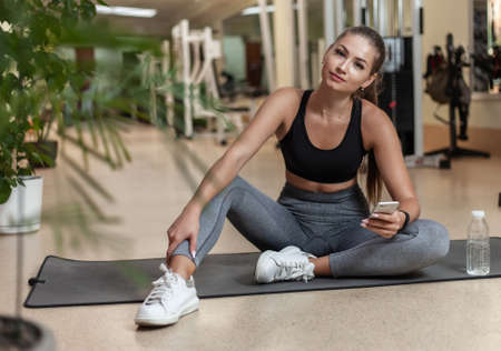 Young sport woman in sportswear using smartphone while sitting on mat in the gym. Workout break. Healthy lifestyle conceptの写真素材