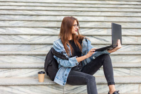 Young modern woman student in denim jacket and glasses sitting on the stairs with laptop. Watching...
