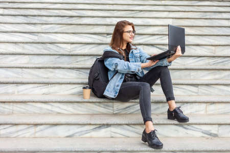 Young modern woman student in denim jacket and glasses sitting on the stairs with laptop. Watching video. Distance learning. Modern youth concept.の写真素材