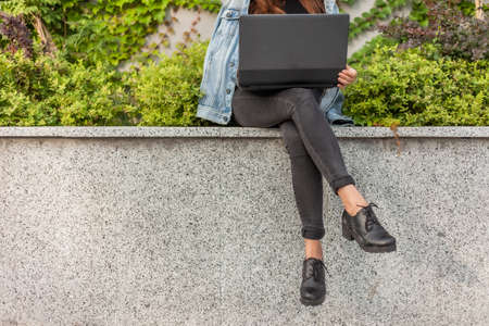 Happy student woman in denim jacket and glasses uses laptop while sitting in the city park. Crop...