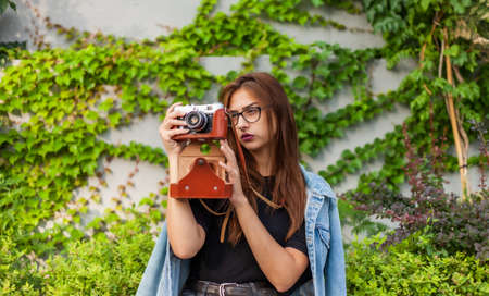 Young hipster woman in denim jacket and glasses enjoys film retro camera outdoors