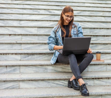 Young modern woman student in a denim jacket sitting on the stairs and watching video on laptop. Distance learning. Modern youth concept.の写真素材