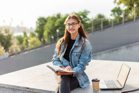 Young attractive student woman is reading a book while sitting on the stairs in the city. The...