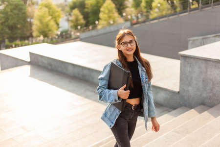 Young student woman in a denim jacket and glasses climbs the stairs with a laptop in her hands in the cityの写真素材
