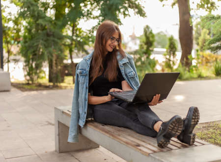 Online call. Distance learning. A young woman student in a denim jacket and glasses looks at a laptop screen while sitting on bench in parkの写真素材