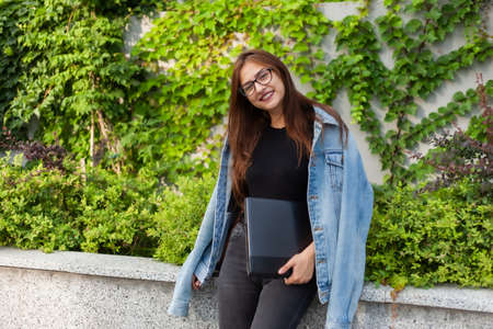 Happy student woman in denim jacket and glasses with laptop in her hands in the city