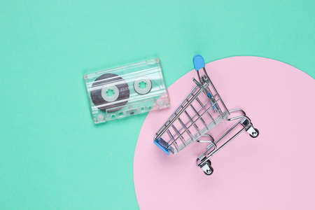 Mini shopping trolley with retro audio cassette on blue background with pink pastel circle. Pop...