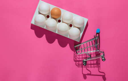 Cart for shopping and chicken eggs in a tray on pink background. Minimalism food concept. Top viewの写真素材