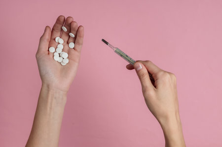 Female hands hold pills and thermometer on a pink background. Medical concept, treatment for colds....