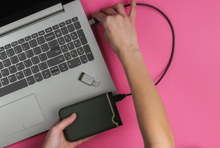 Female hand connects external to a laptop on pink paper background. Top view