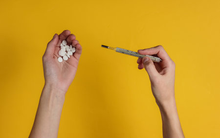 Female hands hold pills and thermometer on yellow background. Medical concept, treatment for colds....