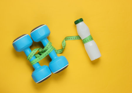 Minimalistic diet and sport concept. Dumbbells, bottle of kefir, measuring tape on yellow paper background. Top viewの写真素材