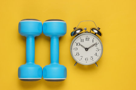 Training time! Dumbbell, alarm clock on yellow background. Minimalistic sport concept. Top viewの写真素材