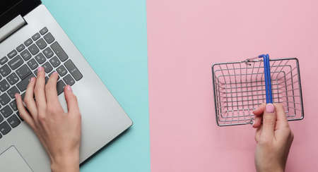Online shopping concept. Female hands hold mini shopping basket and type on laptop keyboard on...