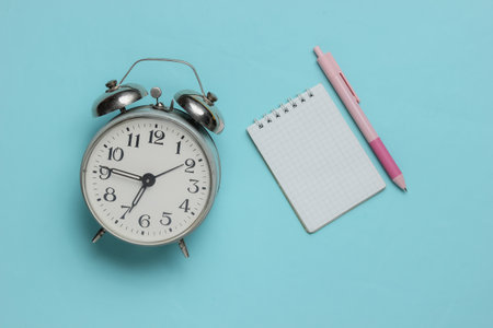 Alarm clock and pink pen with notebook close-up on blue background. Pastel color trend