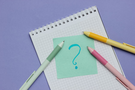 Notepad with pens and memo piece of paper with question mark on purple background. Top view