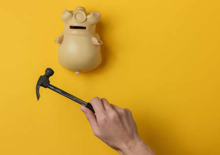 Female hand smashes a piggy bank with a hammer on yellow background. Minimalistic concept. Top view